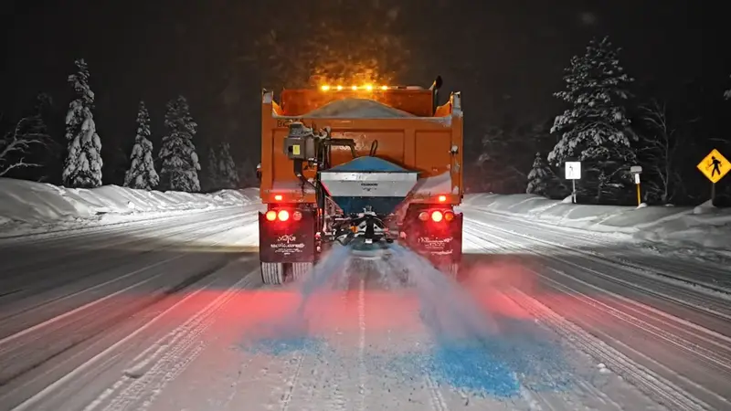 Orange spreader truck throwing treated salt on a snowy road at night, seen from behind